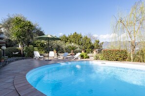 Une piscine entourée de verdure et d’un mur de pierre, avec des chaises longues et un parasol.