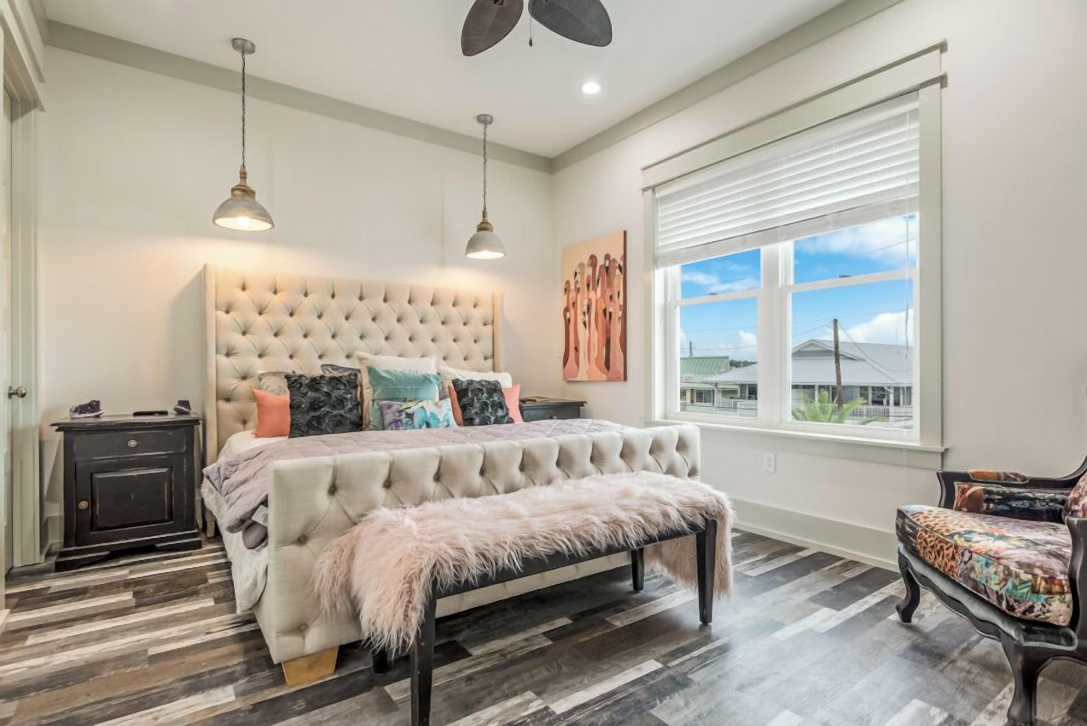 A bedroom with a tufted headboard, a bed with multiple pillows, a bench, a nightstand, and a window with blinds.