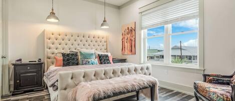 A bedroom with a tufted headboard, a bed with multiple pillows, a bench, a nightstand, and a window with blinds.