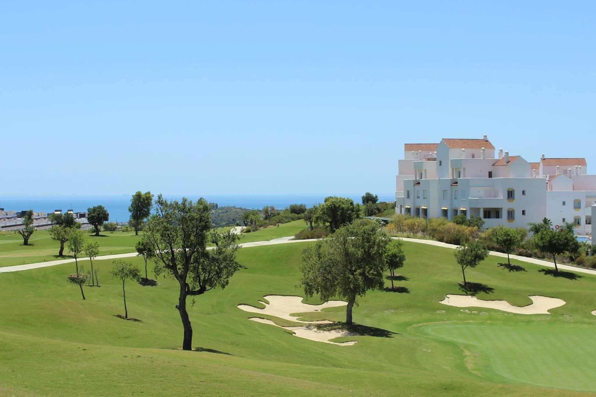 En golfbane med sandbunker og træer, en klar blå himmel og en bygning med rødt tegltag i baggrunden.