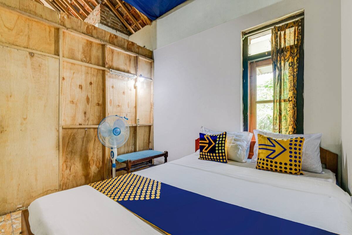 Une pièce avec un lit, un mur en bois, une fenêtre avec des rideaux et un ventilateur.