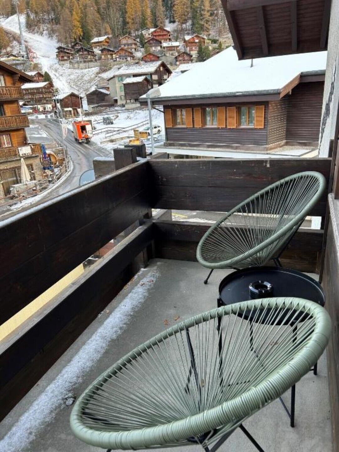 A balcony with two green wire chairs and a small round table, overlooking a snowy village with wooden houses and a road.