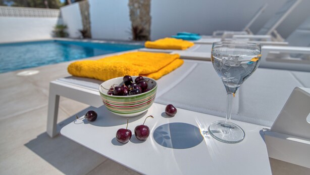 Une scène au bord de la piscine, avec une table sur laquelle se trouvent un bol de cerises, un verre d’eau et des serviettes jaunes.