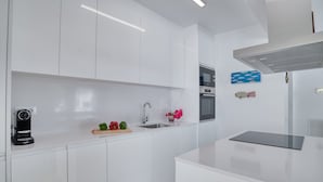 Une cuisine moderne avec des meubles blancs, un four encastré, une machine à café et un petit plan de travail agrémenté d’une planche à découper et de quelques légumes.