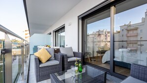 Een balkon met rieten meubels, een glazen tafel met flessen en uitzicht op gebouwen.
