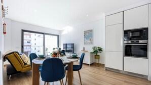 Een moderne keuken met inbouwapparatuur, een zithoek met een gele bank en blauwe stoelen, en een groot raam met uitzicht op de stad.