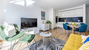 Een moderne woonkamer met een flatscreen televisie, groene stoelen, een gele bank en een keuken met witte kasten en blauwe stoelen.