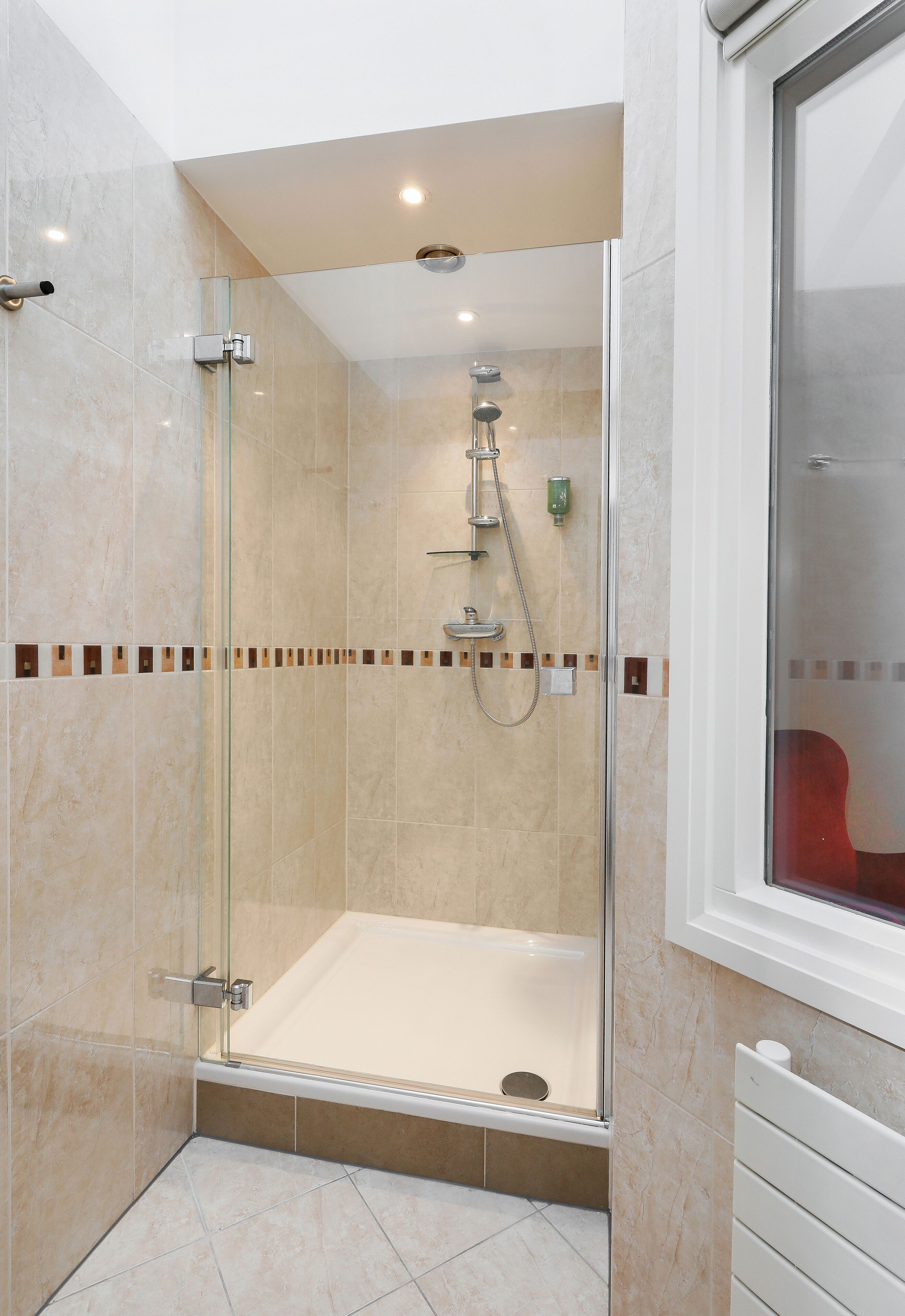 Ein Duschbereich mit Glaswand, Badewanne, geflieste Wände und einem Fenster, durch das ein roter Stuhl zu sehen ist.