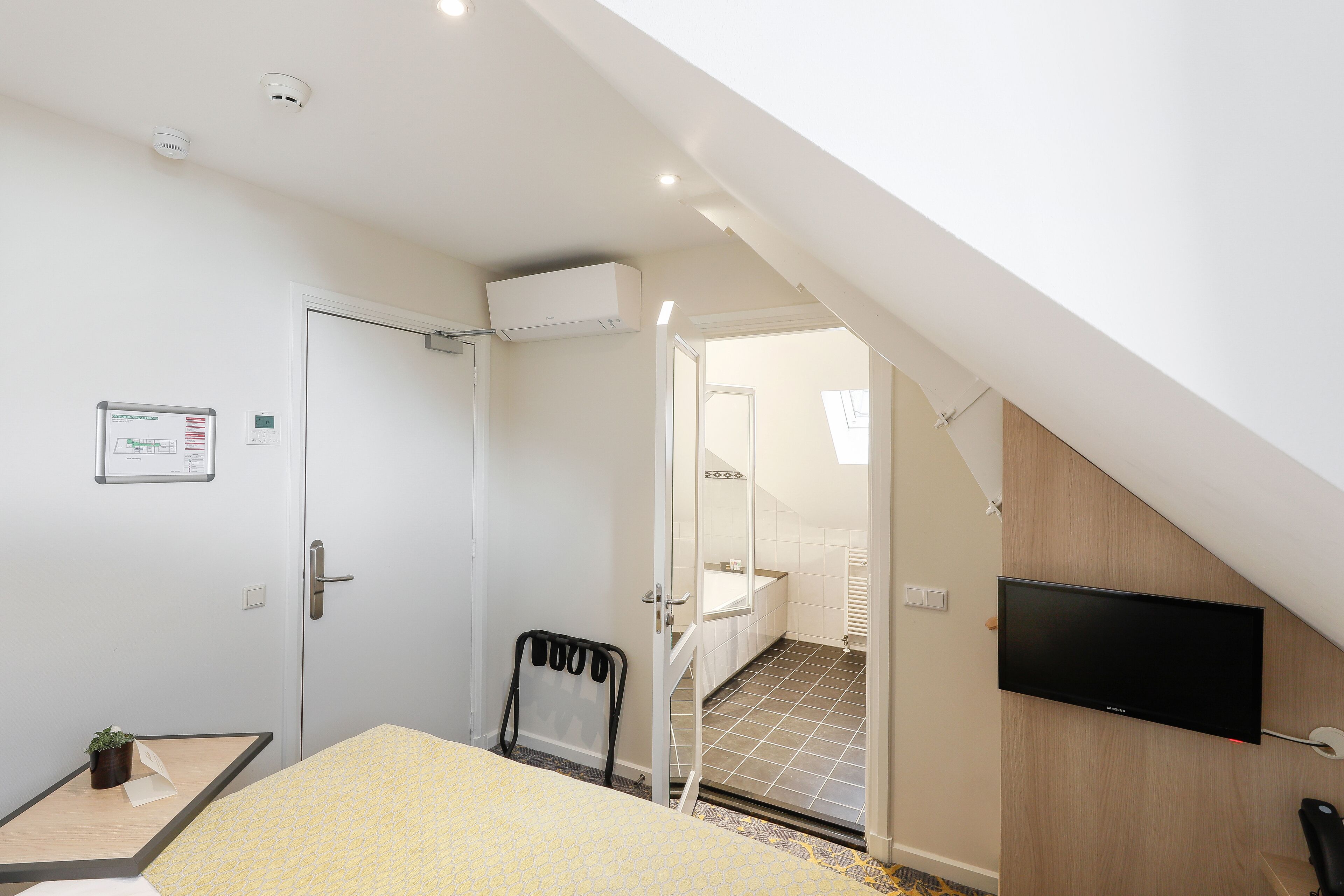 A modern hotel room with a bed, a TV, a small table, and a bathroom visible through an open door.