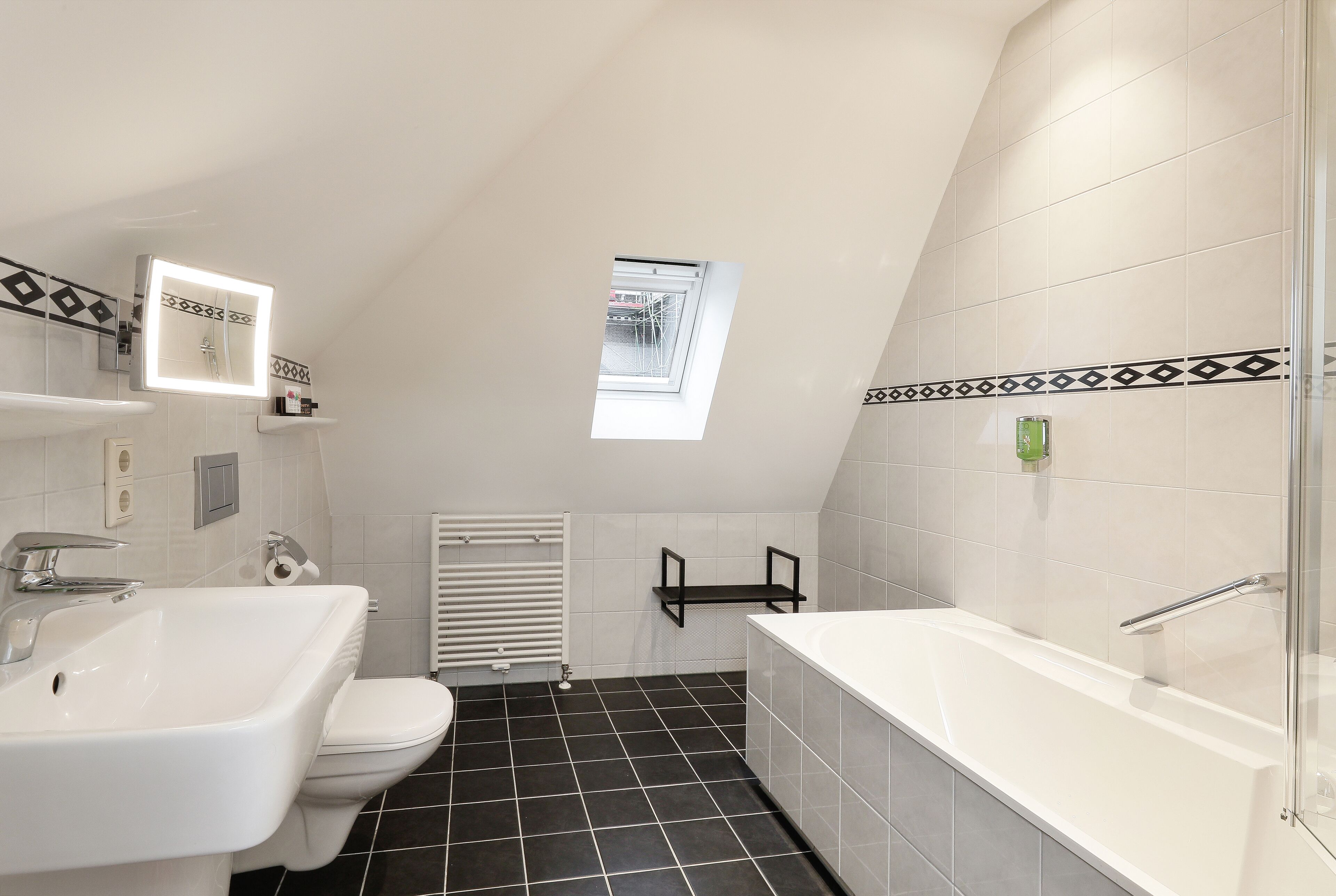 A modern bathroom with a white sink, toilet, and bathtub, black tiled floor, and a skylight.
