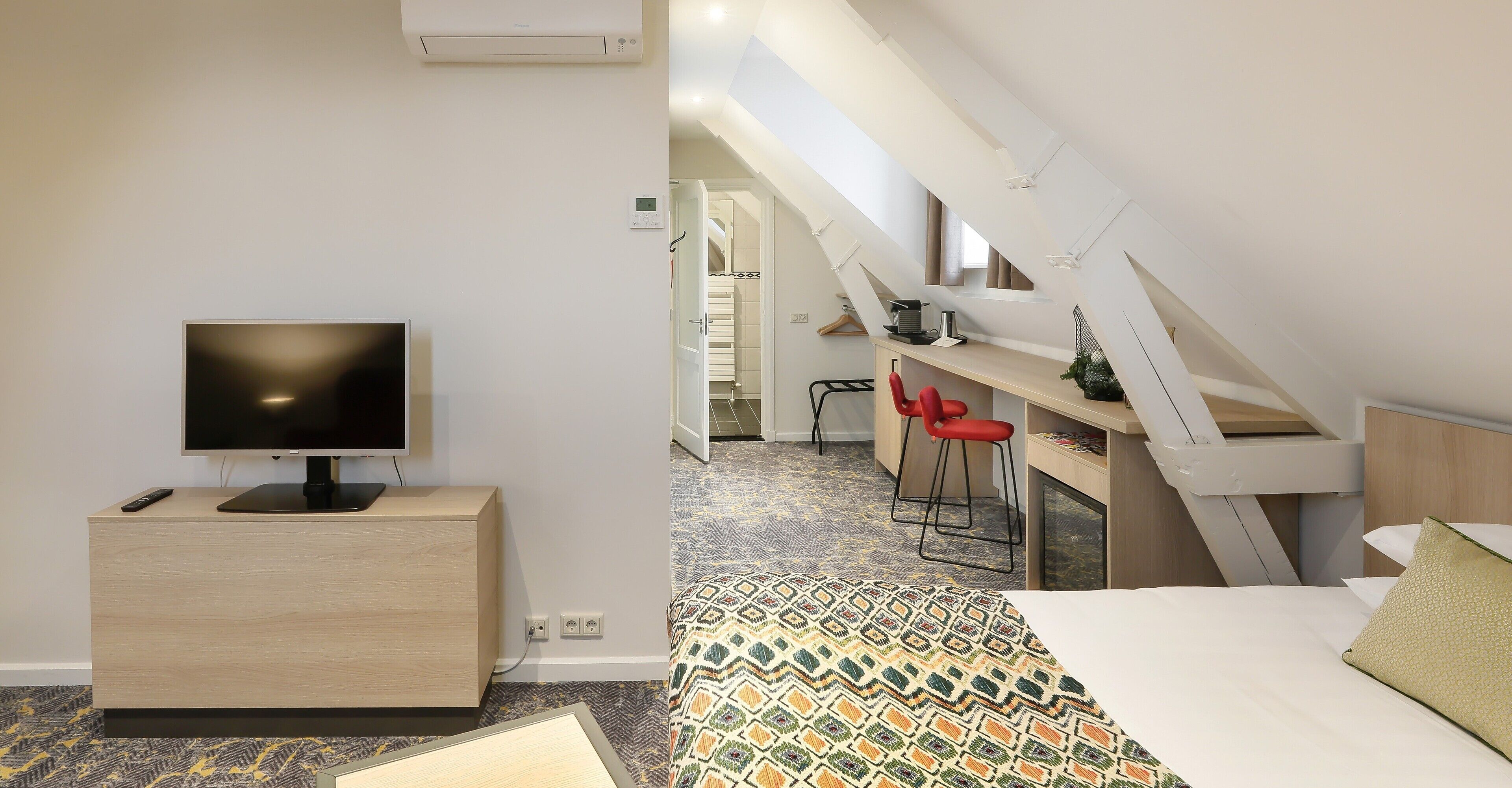Ein modernes Hotelzimmer mit Fernseher auf einer Holzkabine, einer gesteppten Bettdecke und einem Schreibtisch mit Stuhl in der Ecke.