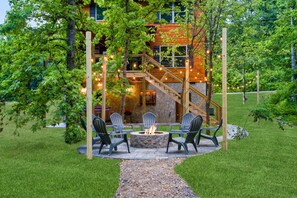 Un espace extérieur accueillant, aménagé avec un foyer, entouré de chaises, devant un bâtiment en bois doté d’un escalier.
