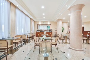 A hotel dining area with glass tables, wicker chairs, and a central buffet.