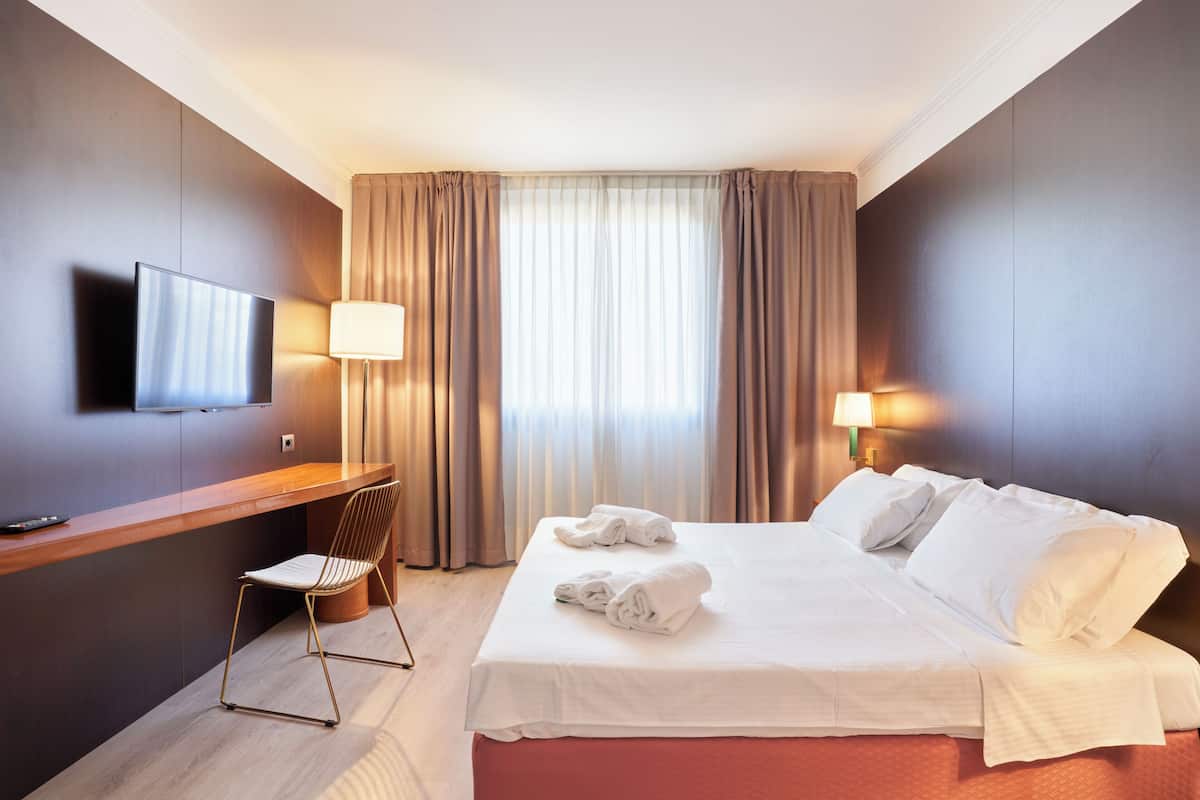 A hotel room with a bed, a desk, a chair, a TV, and curtains.