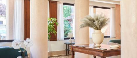 A lobby with a glass coffee table, a green ottoman, a wooden side table with a vase, and large columns.