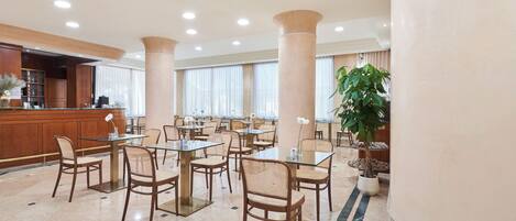 Un amplio vestíbulo de hotel con varias mesas y sillas de comedor, una recepción y grandes pilares.