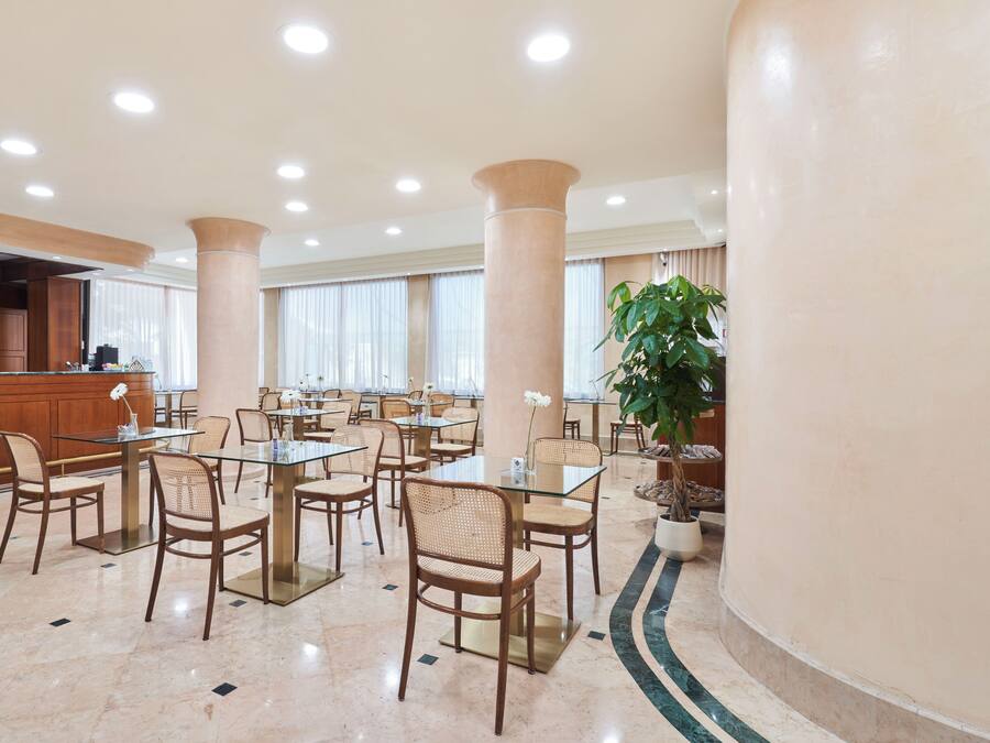 Un amplio vestíbulo de hotel con varias mesas y sillas de comedor, una recepción y grandes pilares.