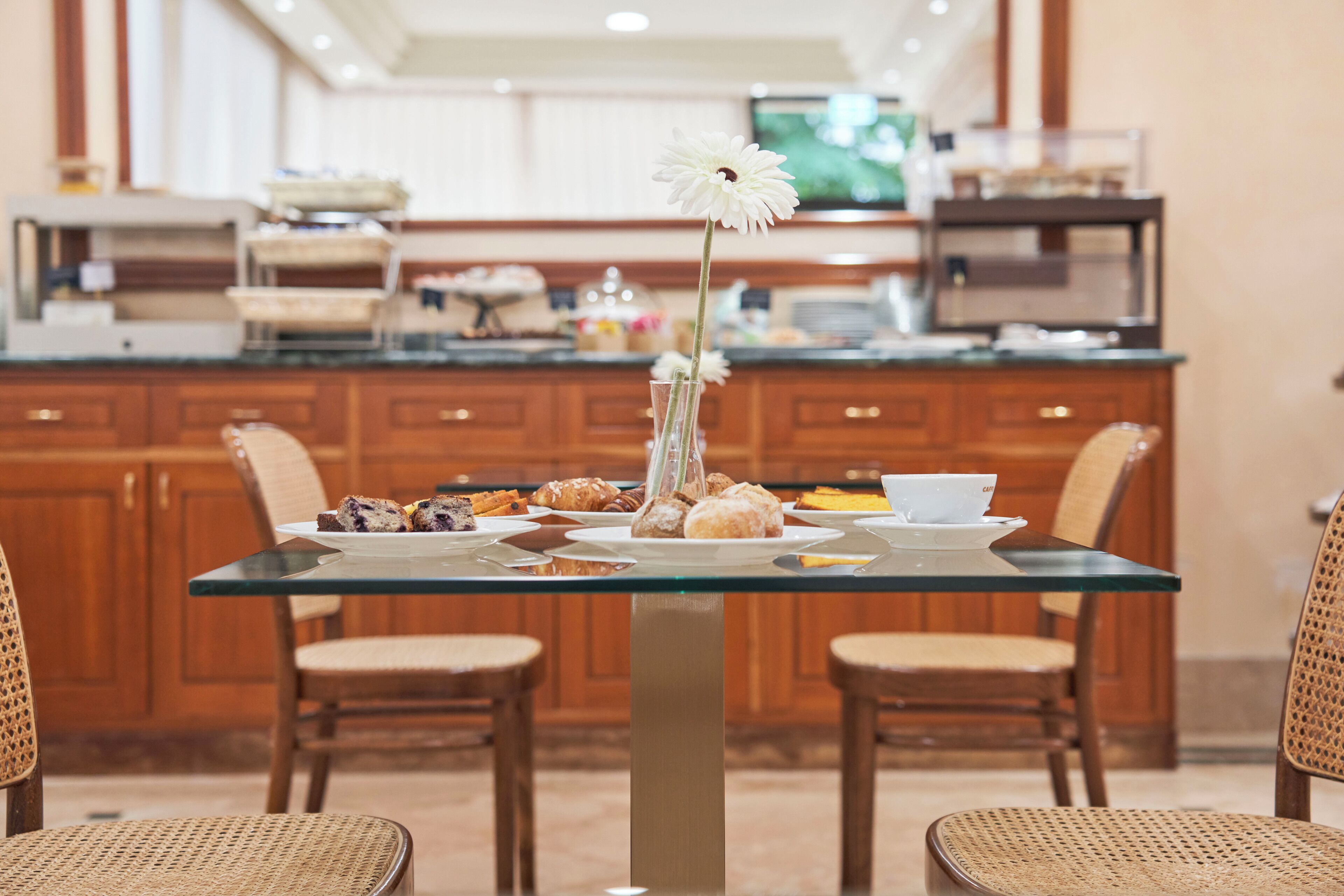 A breakfast setting with a glass table, wicker chairs, and a variety of pastries and a white flower in a vase.