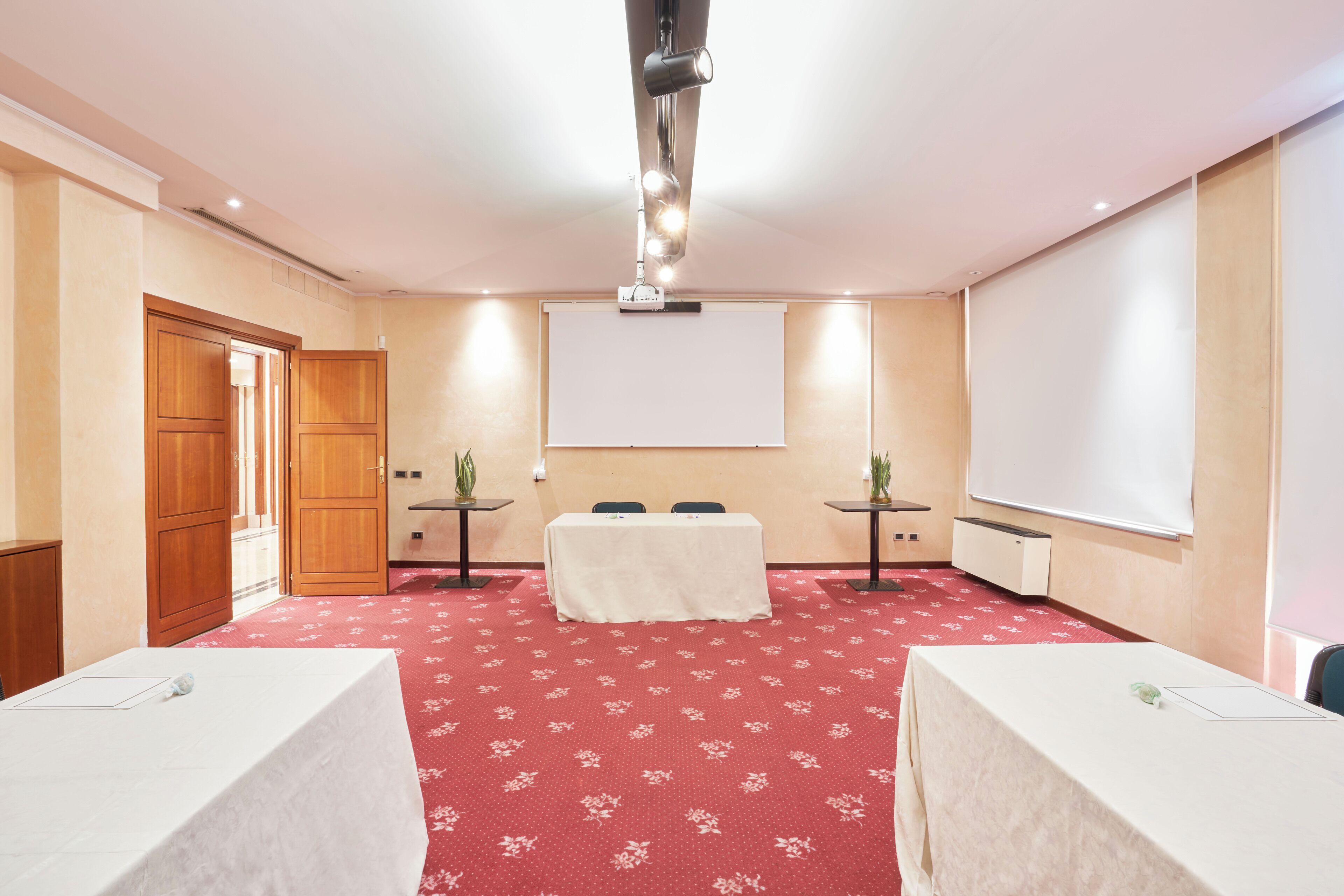 A conference room with a red carpet, a white projection screen, two tables with white tablecloths, and a wooden door.