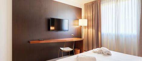 A hotel room with a bed, a TV mounted on the wall, a chair, a lamp, and a window with curtains.