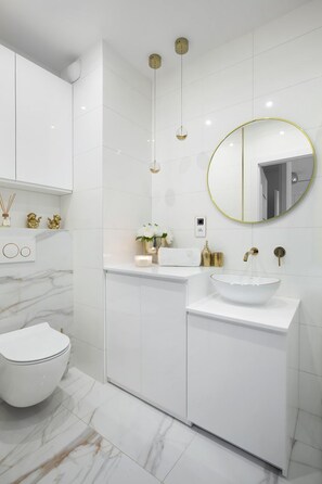 A modern bathroom with a white toilet, a white sink with a gold faucet, and a round mirror with a gold frame. - Elite Apartments Letnica Glamour (Gdansk)