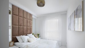 Une chambre à coucher moderne avec une tête de lit capitonnée, un grand lit avec une literie blanche, une table de chevet avec une lampe et une grande fenêtre ornée de rideaux légers.