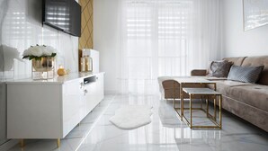Un salon moderne avec un canapé blanc, une table basse, un téléviseur fixé au mur et une grande fenêtre ornée de rideaux légers.
