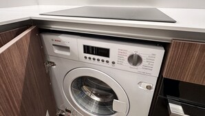 Una lavatrice compatta e moderna con display digitale e pannello di controllo, posizionata in un angolo di una stanza con mobili in legno e un piano di lavoro bianco.