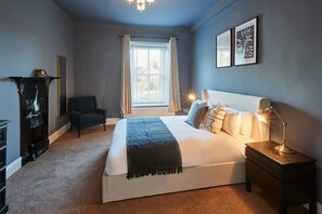 Une chambre à coucher avec un grand lit, une table de chevet avec une lampe, une chaise, une cheminée et une fenêtre avec des rideaux.