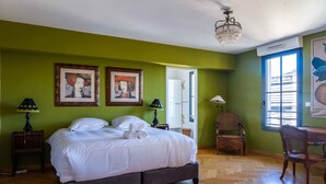 A bedroom with a large bed, two wall-mounted paintings, a chandelier, a desk with a chair, and a window with a view. - Voltaire - Magnificent 4-bedroom duplex in Triangle d'Or (Bordeaux)