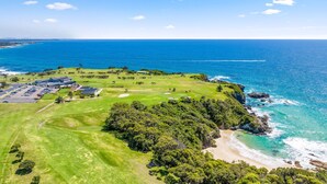 Vista aérea de um campo de golfe costeiro com um clube, estacionamento e vista para o oceano.