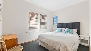 Quarto com cama, duas mesas de cabeceira, um baú de madeira e duas janelas com persianas.