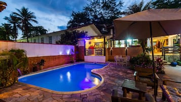 Una piscina con solárium de piedra, rodeada por un patio con mobiliario exterior y una zona cubierta.