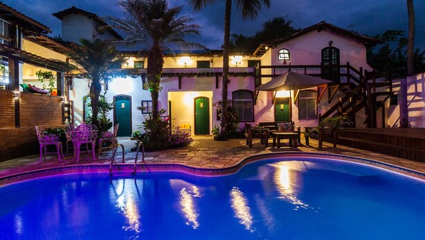 Zona de piscina con borde curvo, rodeada por un patio con sillas y mesas. Hay puertas y ventanas en el edificio, y una escalera que lleva a un nivel superior.