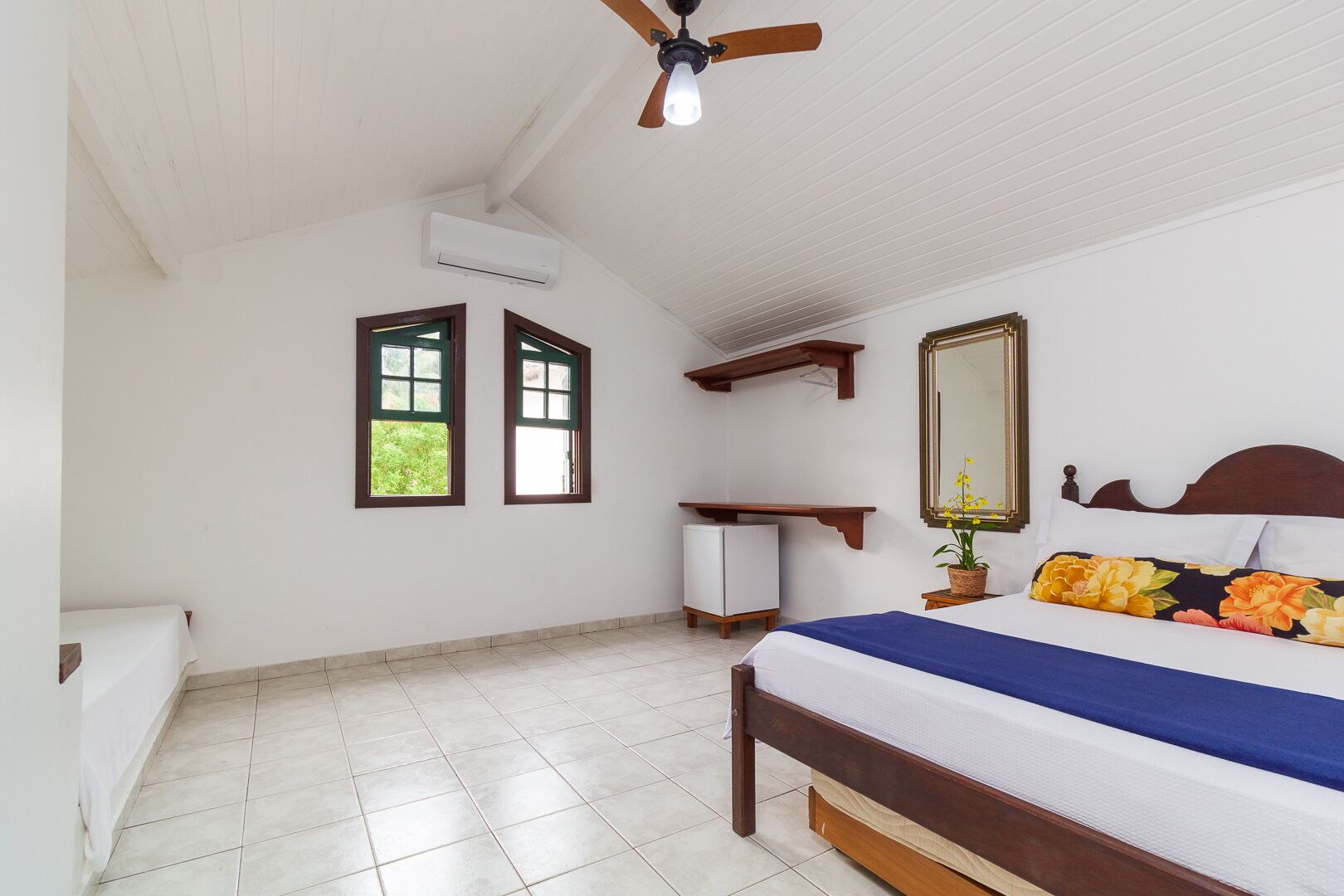 A bedroom with a bed, a ceiling fan, a window, a small white cabinet, and a mirror.