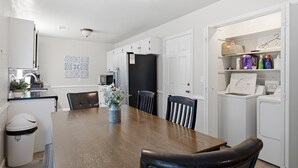 Een moderne keuken met witte kasten, een eettafel en stoelen. Er staat een prullenbak en een magnetron op het aanrecht.