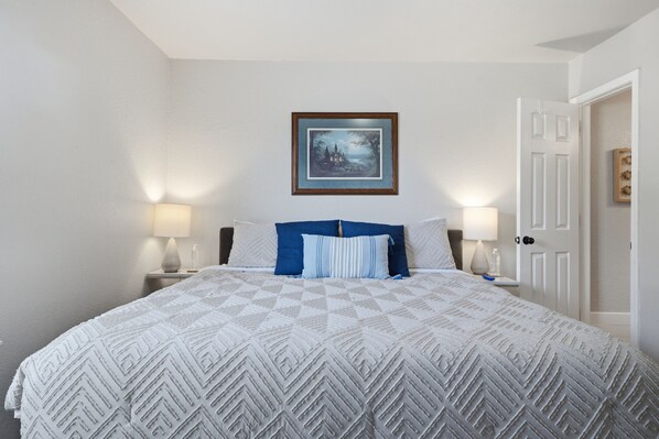 A neatly arranged bedroom with a large bed, two bedside lamps, a framed picture on the wall, and a door to the right. - Cozy 3BR family retreat with a fenced backyard, patio, and everything you need for a comfortable stay in Broken Arrow. Pet friendly! (Broken Arrow)