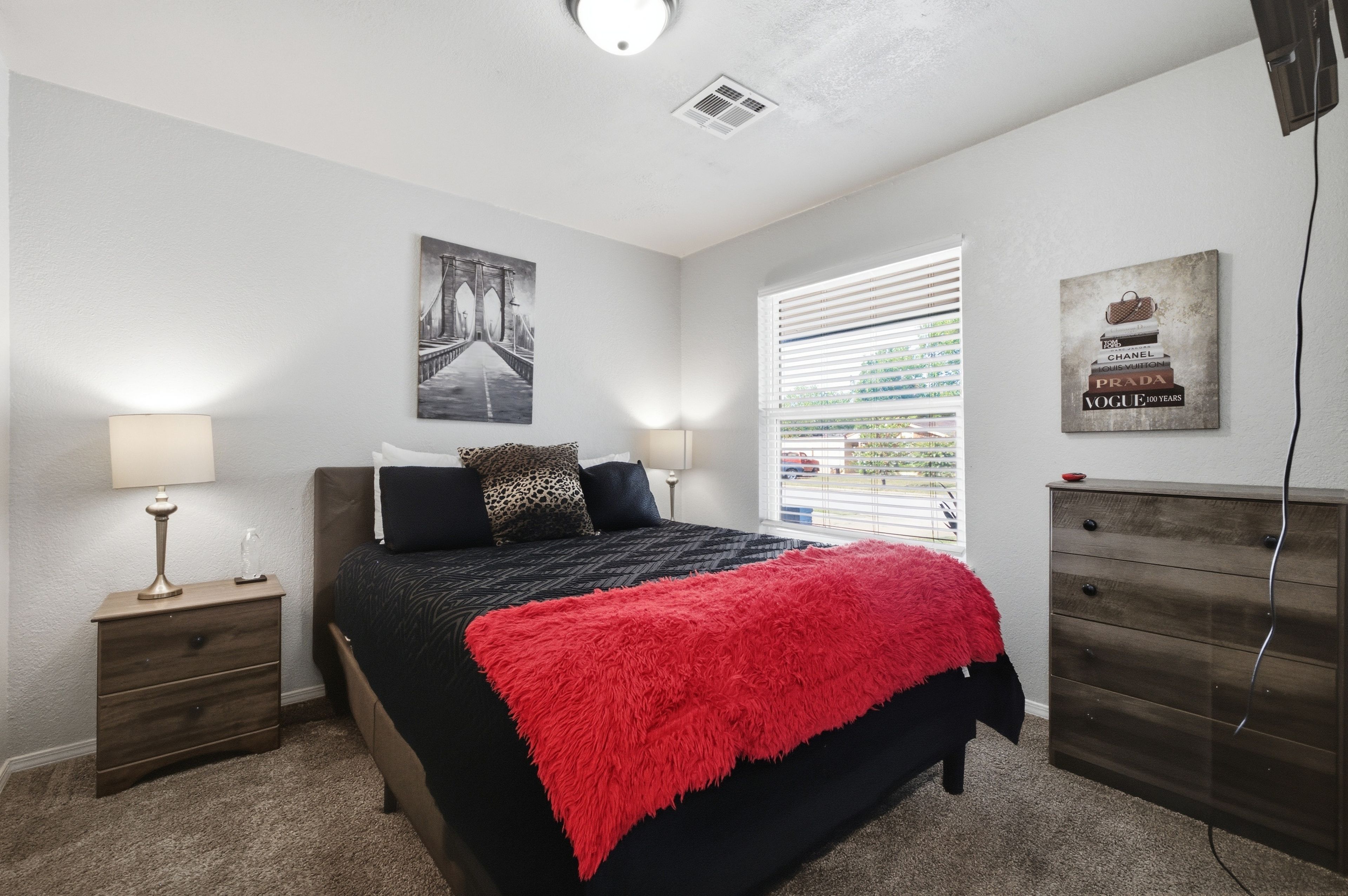 A bedroom with a bed, bedside tables, a window with blinds, a wall-mounted artwork, and a dresser.