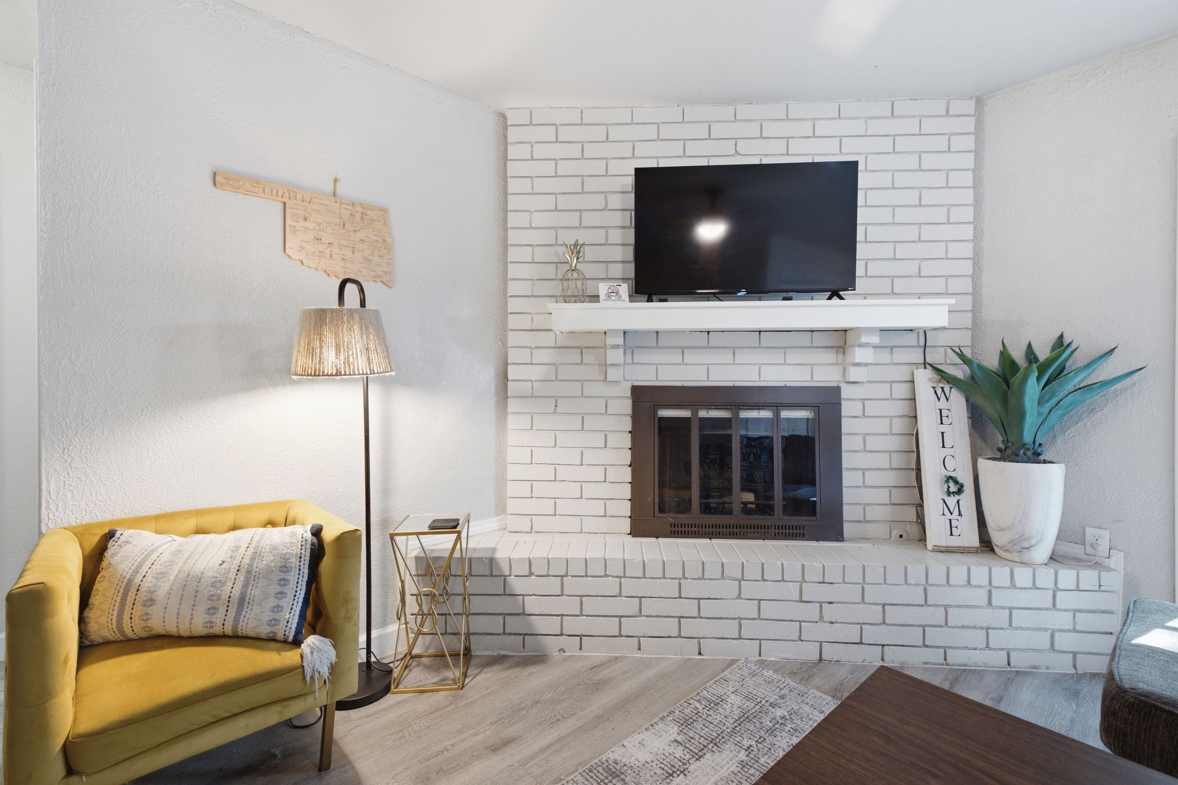 A living room with a white brick fireplace, a flat-screen TV mounted above it, a yellow sofa, and a floor lamp.