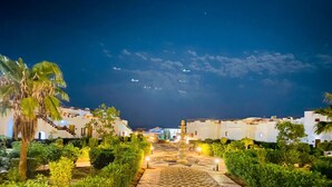 A nighttime view of a courtyard with a patterned pathway leading to a central fountain, surrounded by lush greenery and white buildings with flat roofs. - Grand Halomy resort (Sharm El Sheikh)
