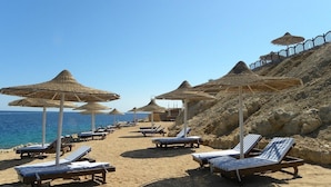 A beach with sun loungers and thatched umbrellas, a rocky hillside, and a clear blue sky. - Grand Halomy resort (Sharm El Sheikh)