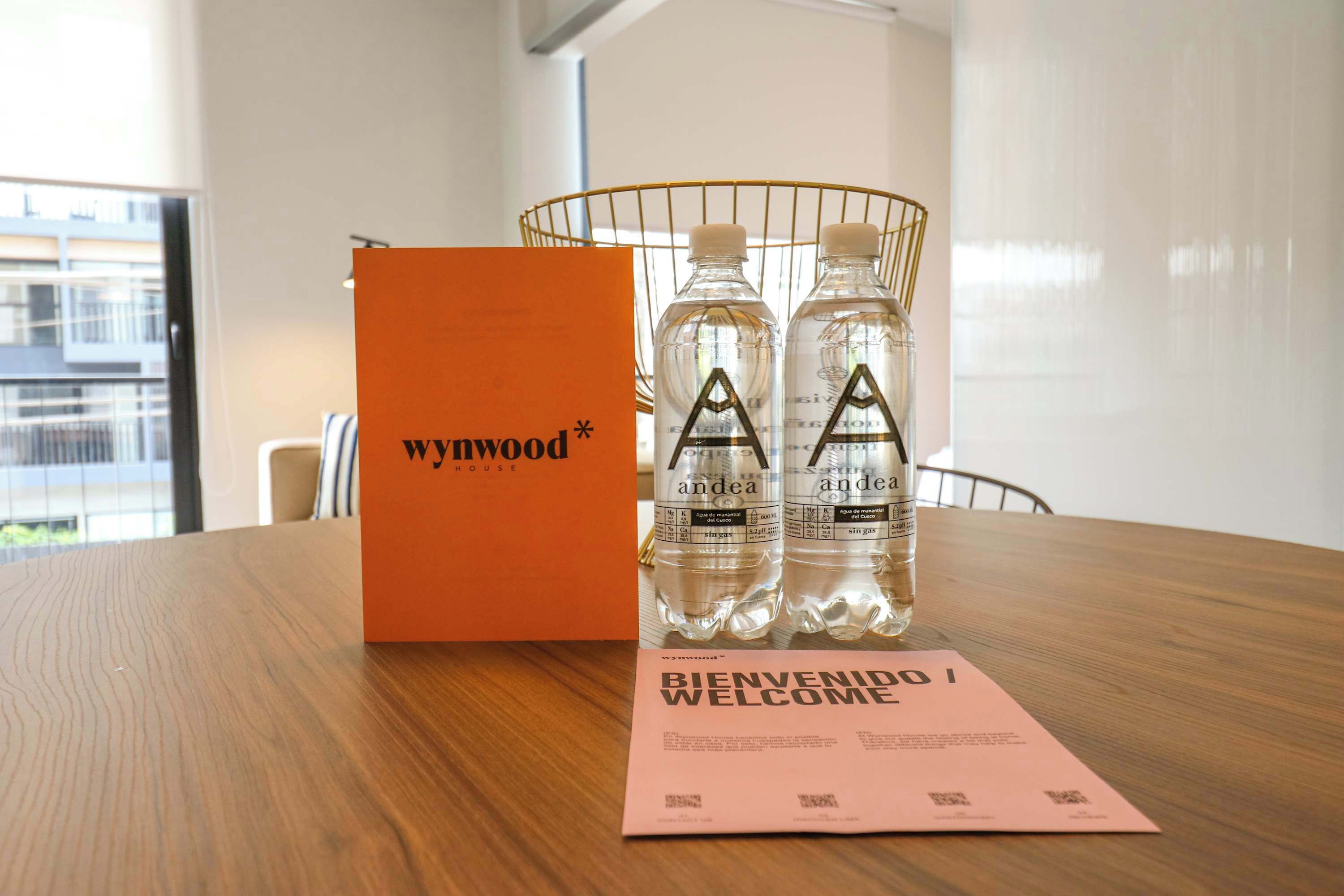 Una mesa de madera con una tarjeta de bienvenida, dos botellas de agua y una caja naranja con el rótulo 'Woodhouse'.