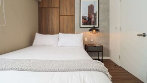 Ein ordentlich bezogenes Bett mit weißen Leinen und einer gemusterten Tagesdecke, eine Wand aus Holzverkleidung, ein gerahmtes Stadtbild und ein kleiner Nachttisch mit Lampe.