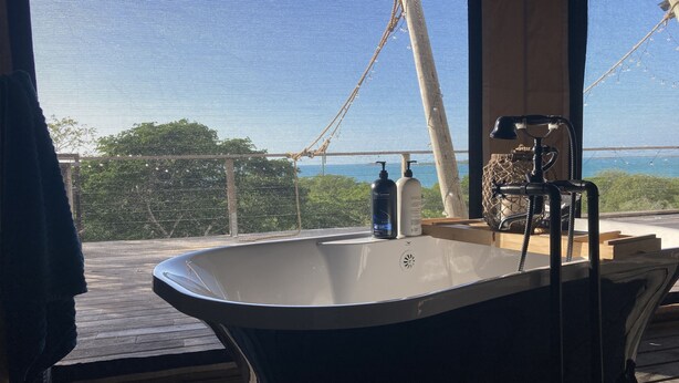 Ein Badezimmer mit freistehender Badewanne, einem Fenster mit Blick auf Grünflächen und ein Gewässer, sowie Pflegeprodukten auf dem Fensterbrett.
