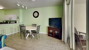 A dining area with a table and chairs, a kitchen with white cabinets, a clock on the wall, and a TV stand with books. - Shores of Panama 524 | Pool, Spa, Gym! (Panama City Beach)