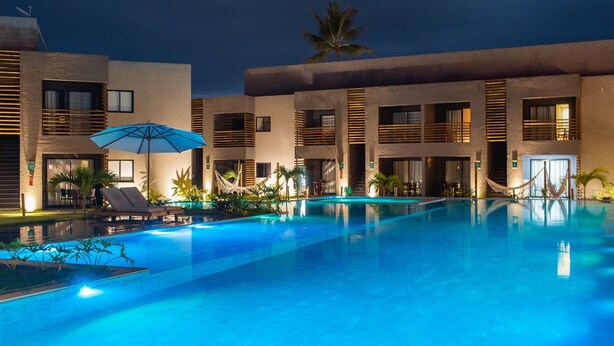 A swimming pool area with lounge chairs and umbrellas, surrounded by buildings with balconies and palm trees. - Flat I 002 Villas Manatee Patacho - AL (Porto de Pedras)