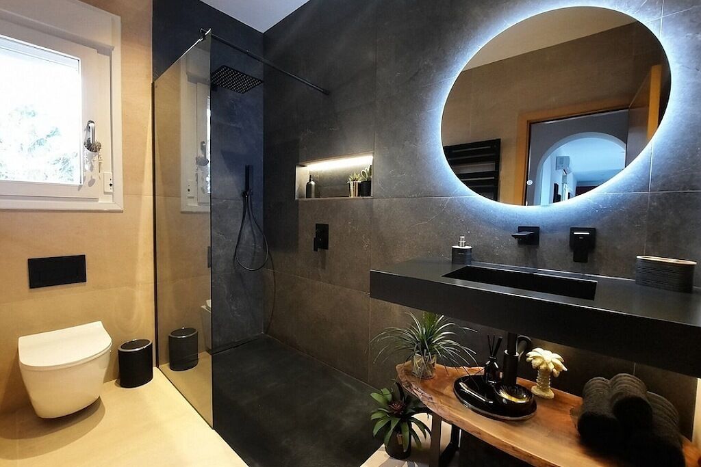 Un baño moderno con un espejo grande, una ducha y una zona de lavabo con plantas y artículos de aseo.