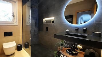 Un baño moderno con un espejo grande, una ducha y una zona de lavabo con plantas y artículos de aseo.
