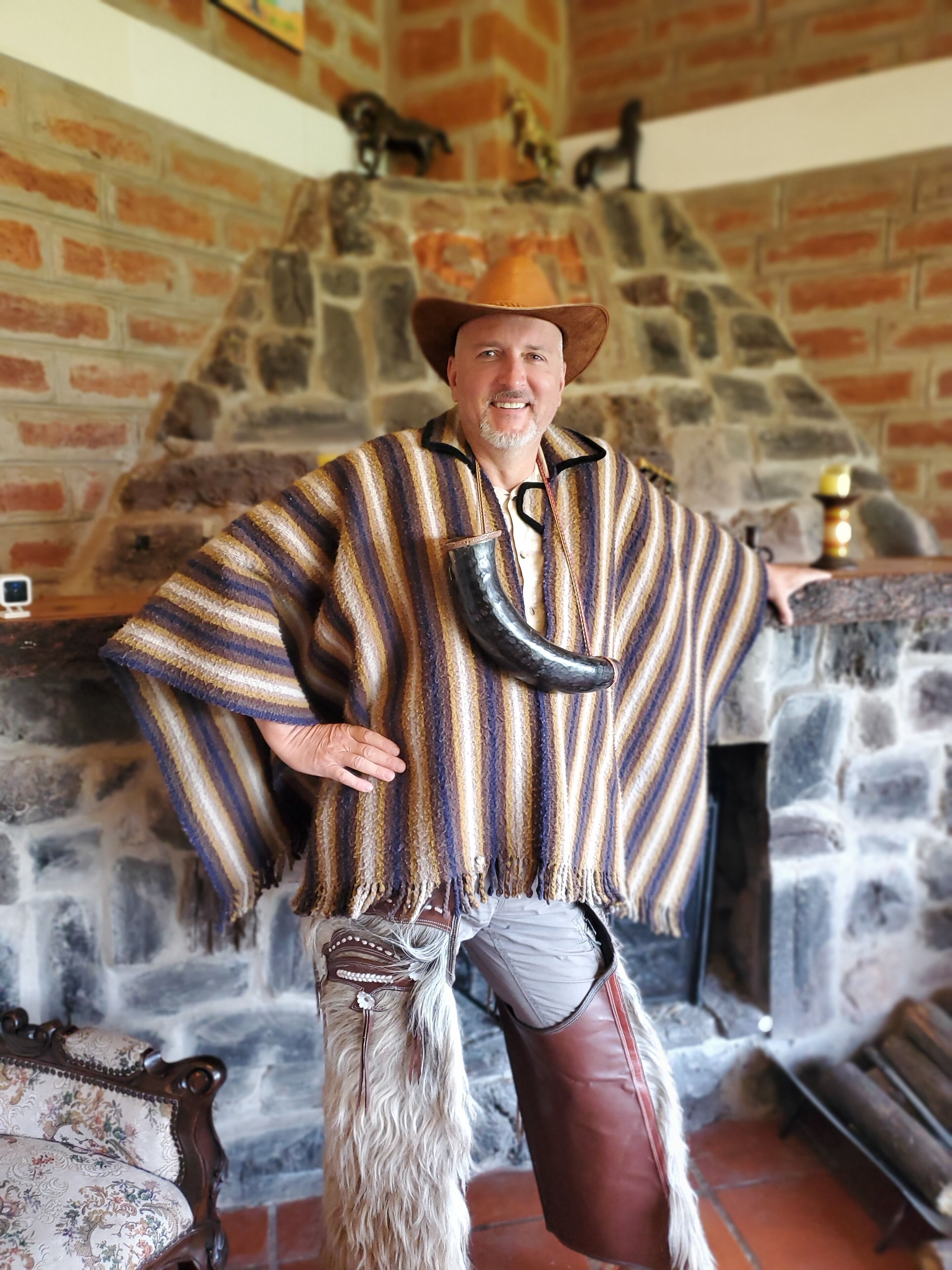 Una persona vestida con traje tradicional frente a una chimenea de piedra, con pared de ladrillo y adornos.