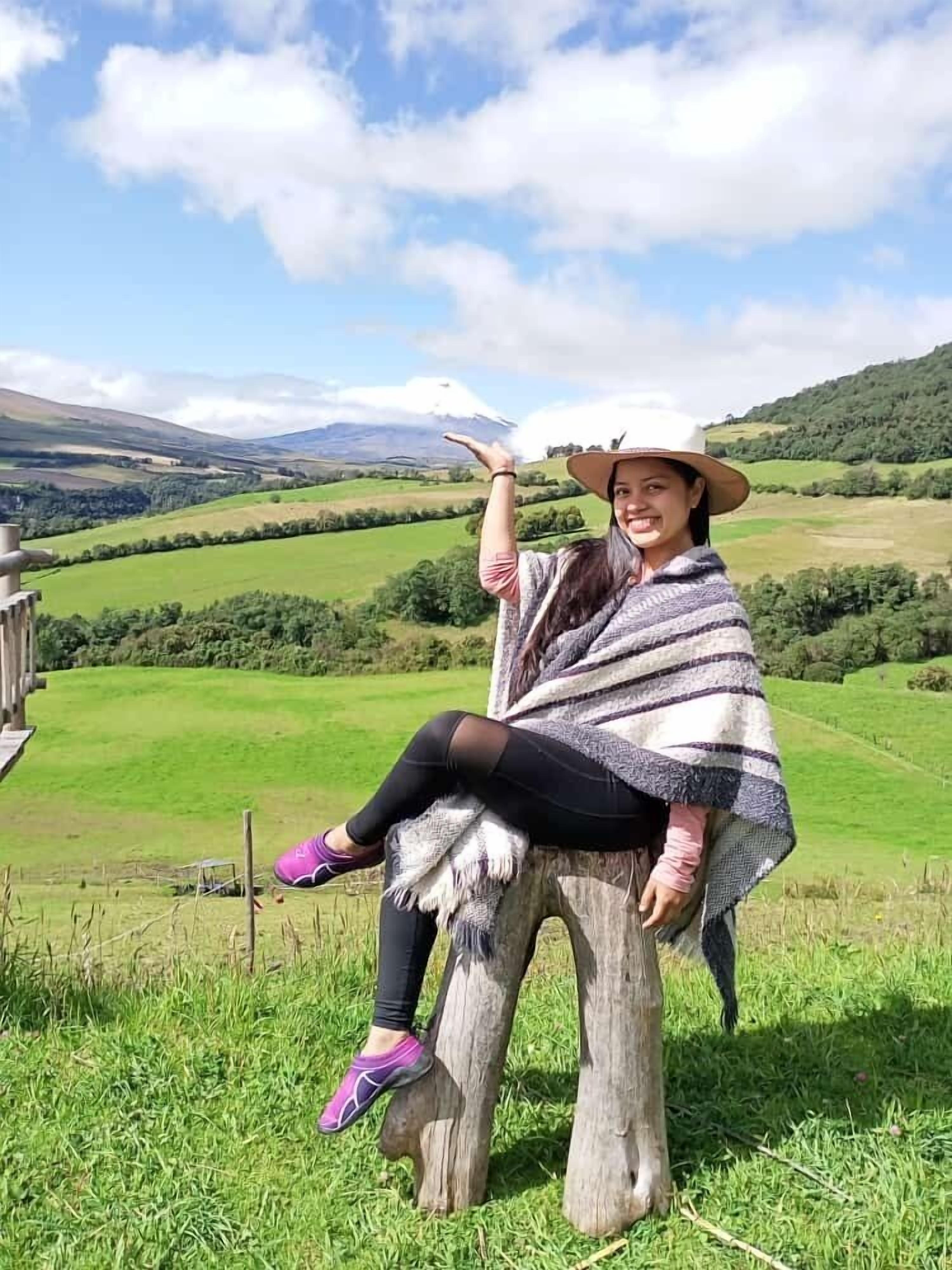 A person sitting on a wooden post in a grassy field with a mountainous landscape in the background.